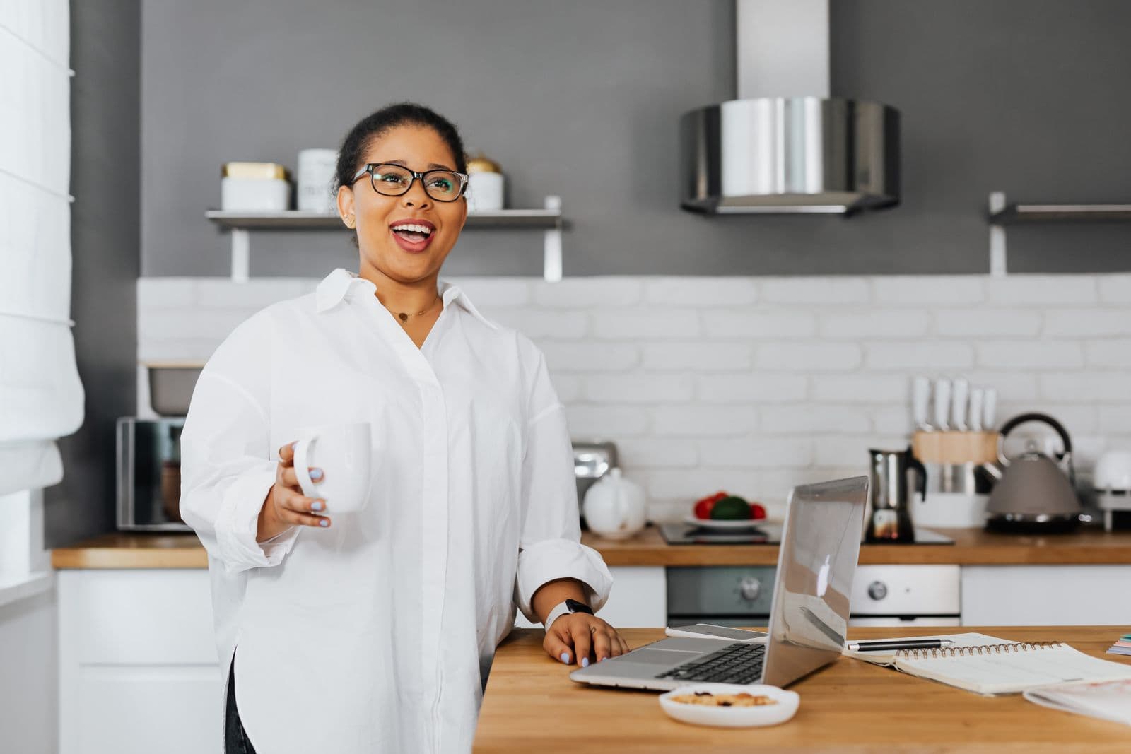 Woman laughing as she stands with a coffee beside her laptop and notebook in a bright kitchen